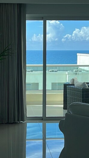 A room with a large window offering a view of the ocean and a building across the water. - Juan Dolio Executive One Bedroom Suite With Ocean Views. (Juan Dolio)