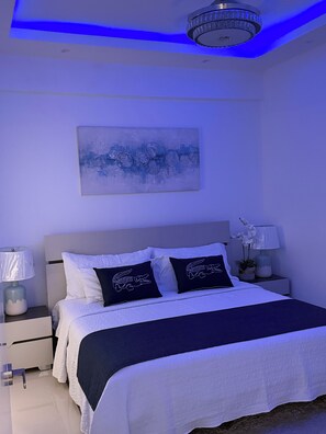 A bedroom with a bed, nightstands, a lamp, a painting, and a ceiling light fixture. - Juan Dolio Executive One Bedroom Suite With Ocean Views. (Juan Dolio)