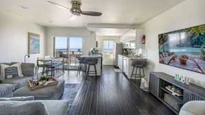 Un salon moderne avec un parquet en bois foncé, un canapé gris, un coin repas avec une table et des chaises hautes, une cuisine avec des meubles blancs et un grand tableau représentant une scène tropicale.
