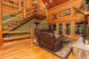 A log cabin interior with a wooden staircase, a leather sofa, and a stone fireplace. - Luxury Lodge in Hocking Hills, 4 Fireplaces, 12-Person Hot Tub, Secret Game Room (Laurelville)