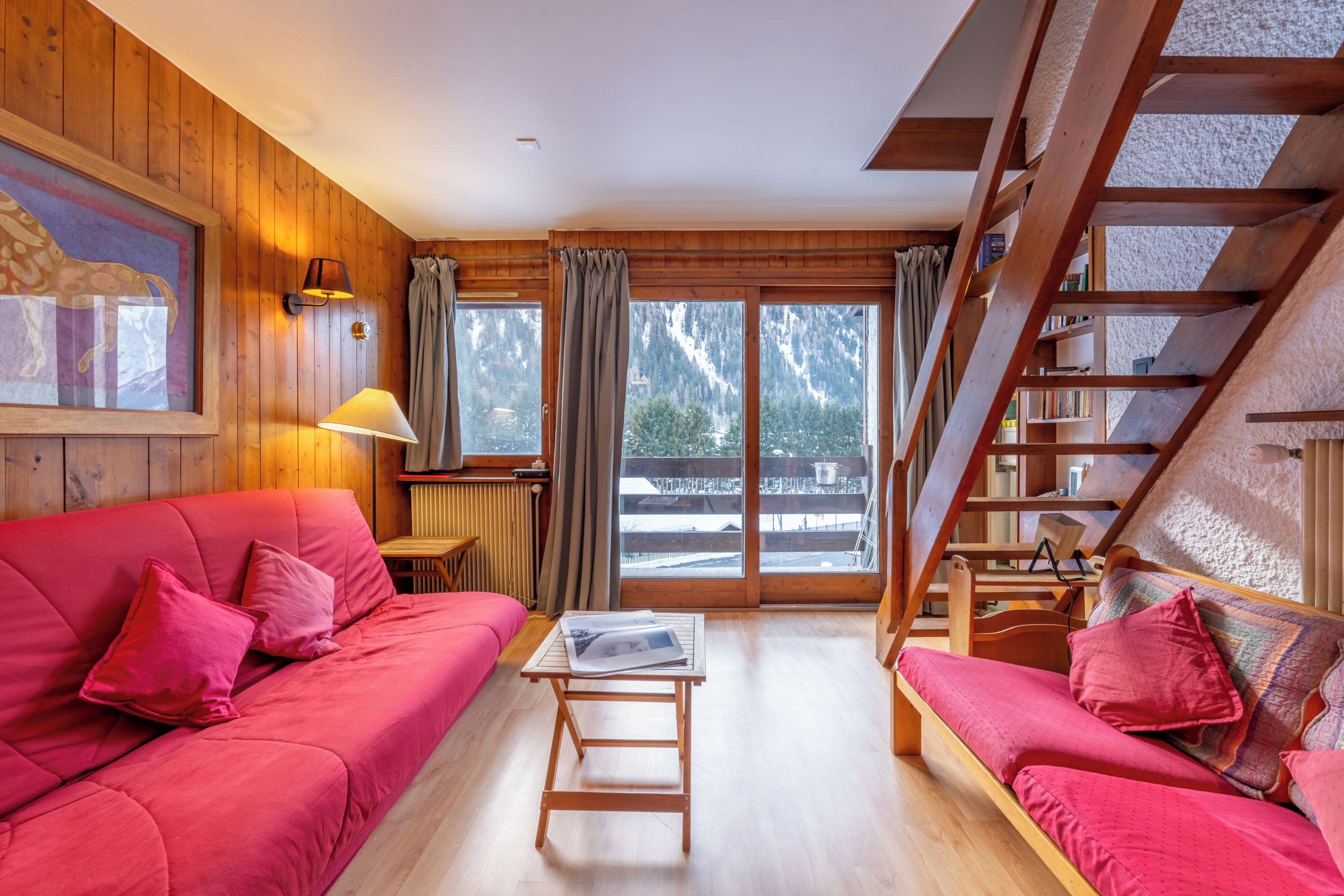 Ein gemütliches Zimmer mit Holzvertäfelung, einem roten Sofa, einem kleinen Tisch und einer Treppe, die auf einen Balkon führt.
