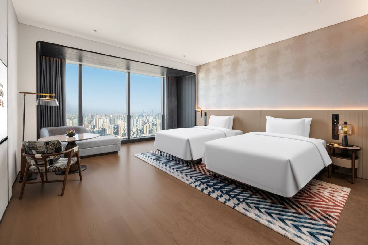 Una habitación de hotel moderna con dos camas, un sofá, una mesa de comedor y un amplio ventanal con vistas a la ciudad.