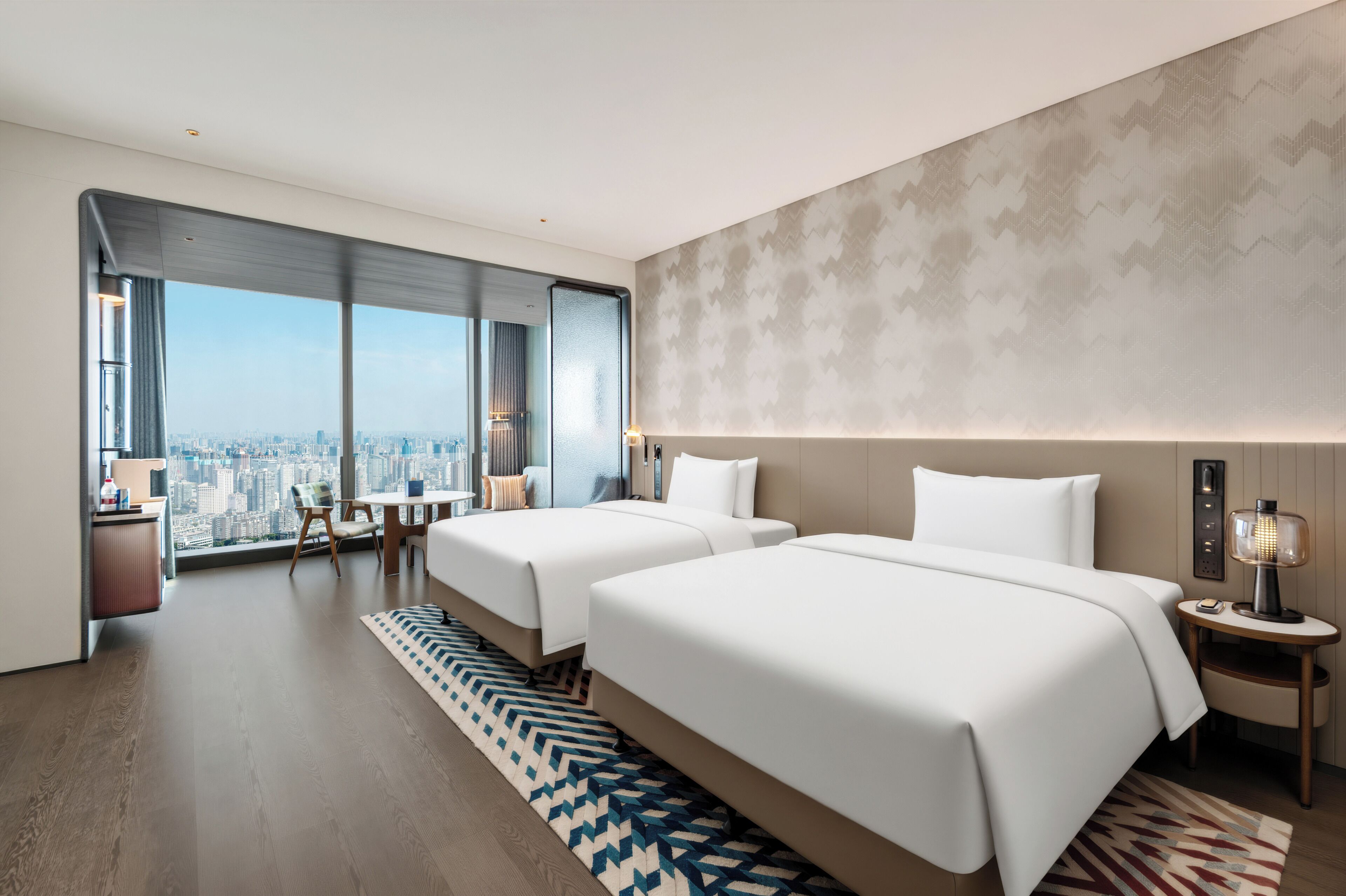 A modern hotel room with two beds, a city view, a desk, and a large window.