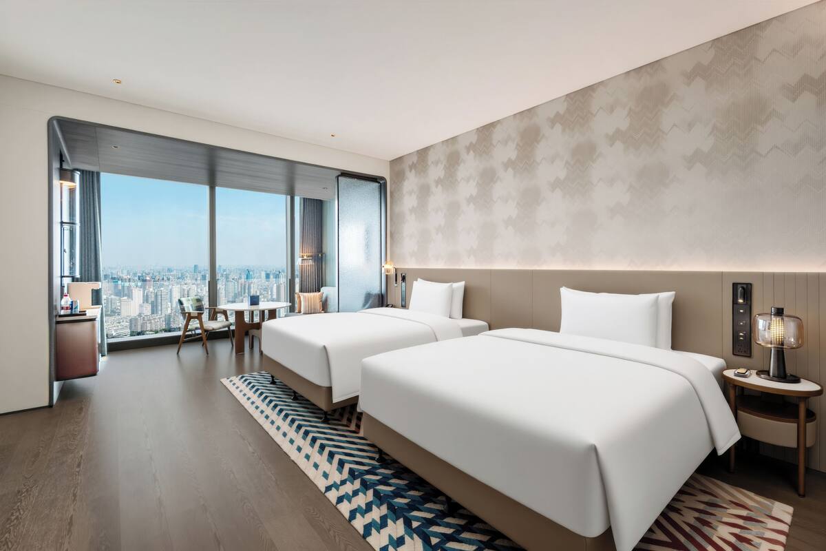 Habitación de hotel moderna con dos camas, vistas a la ciudad, un escritorio y un ventanal grande.