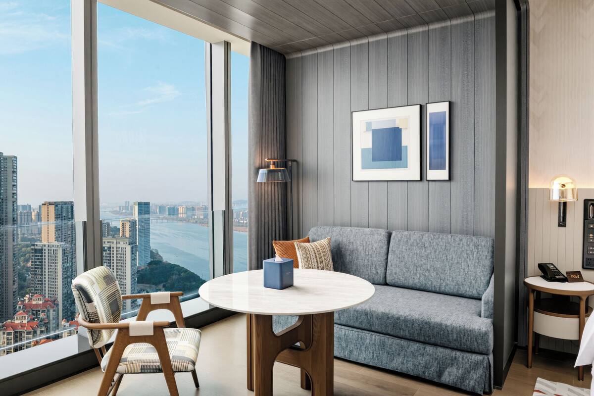 Habitación de hotel moderna con un ventanal grande, una mesa redonda, un sofá y una pequeña mesita auxiliar.