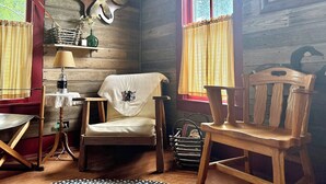 A rustic cabin interior with wooden walls, a chair with a decorative cover, a table with a lamp, and a window with curtains. - Country Farmhouse on a Working Cattle Ranch (El Dorado)