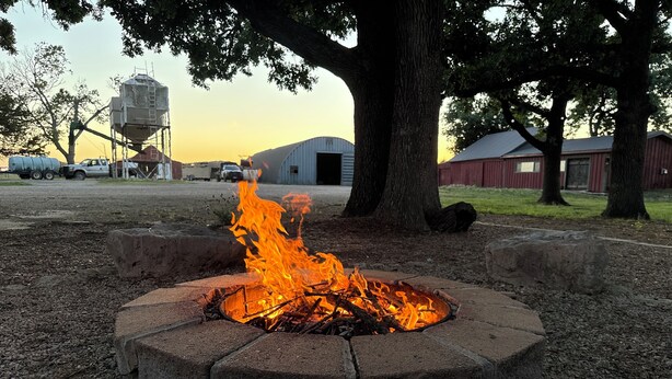 A fire pit in a circular stone design surrounded by gravel, with a tree and buildings in the background. - Country Farmhouse on a Working Cattle Ranch (El Dorado)