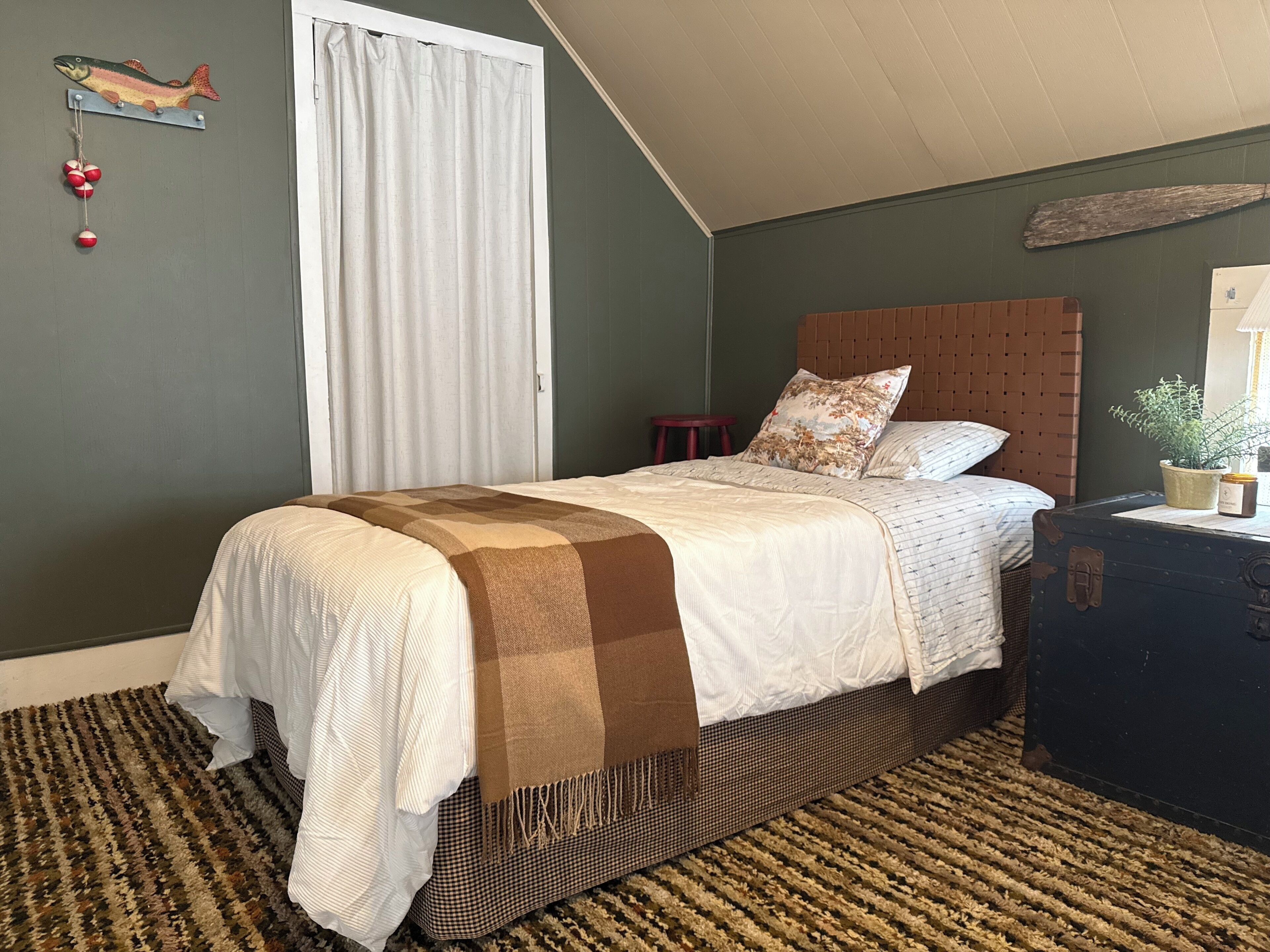 Une chambre à coucher avec un lit, une tête de lit, une table de chevet, une fenêtre avec des rideaux, un élément de décoration fixé au mur et une valise.