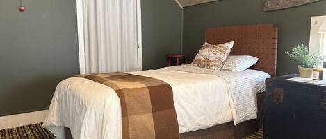 Une chambre à coucher avec un lit, une tête de lit, une table de chevet, une fenêtre avec des rideaux, un élément de décoration fixé au mur et une valise.