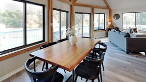 Um quarto amplo com uma mesa de jantar de madeira, cadeiras pretas, um sofá cinzento e grandes janelas com vista para o exterior.