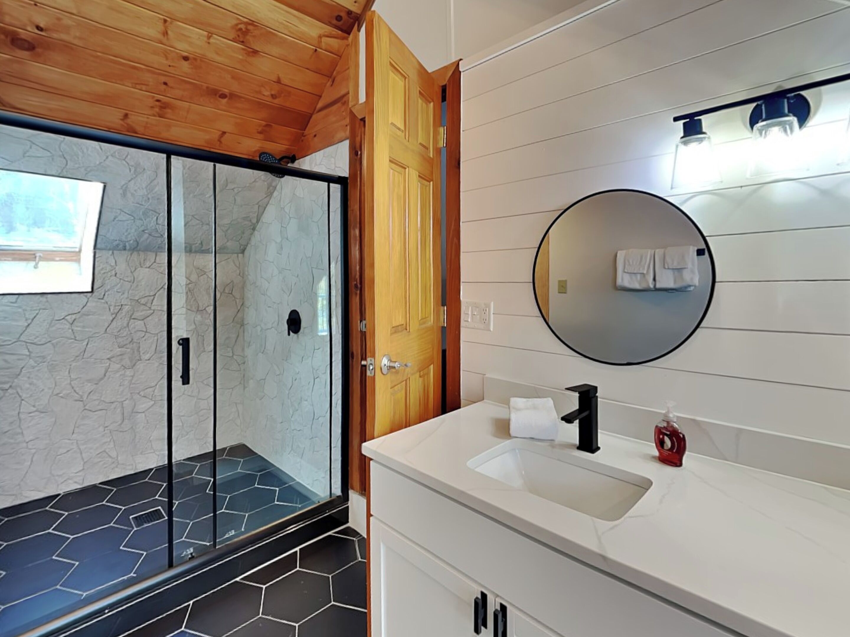 A modern bathroom with a glass shower enclosure, a wooden ceiling, and a white vanity with a round mirror.