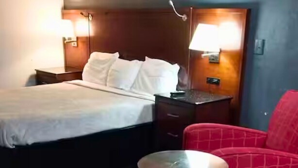Quarto de hotel com cama, mesas de cabeceira, uma mesa redonda e uma cadeira vermelha.