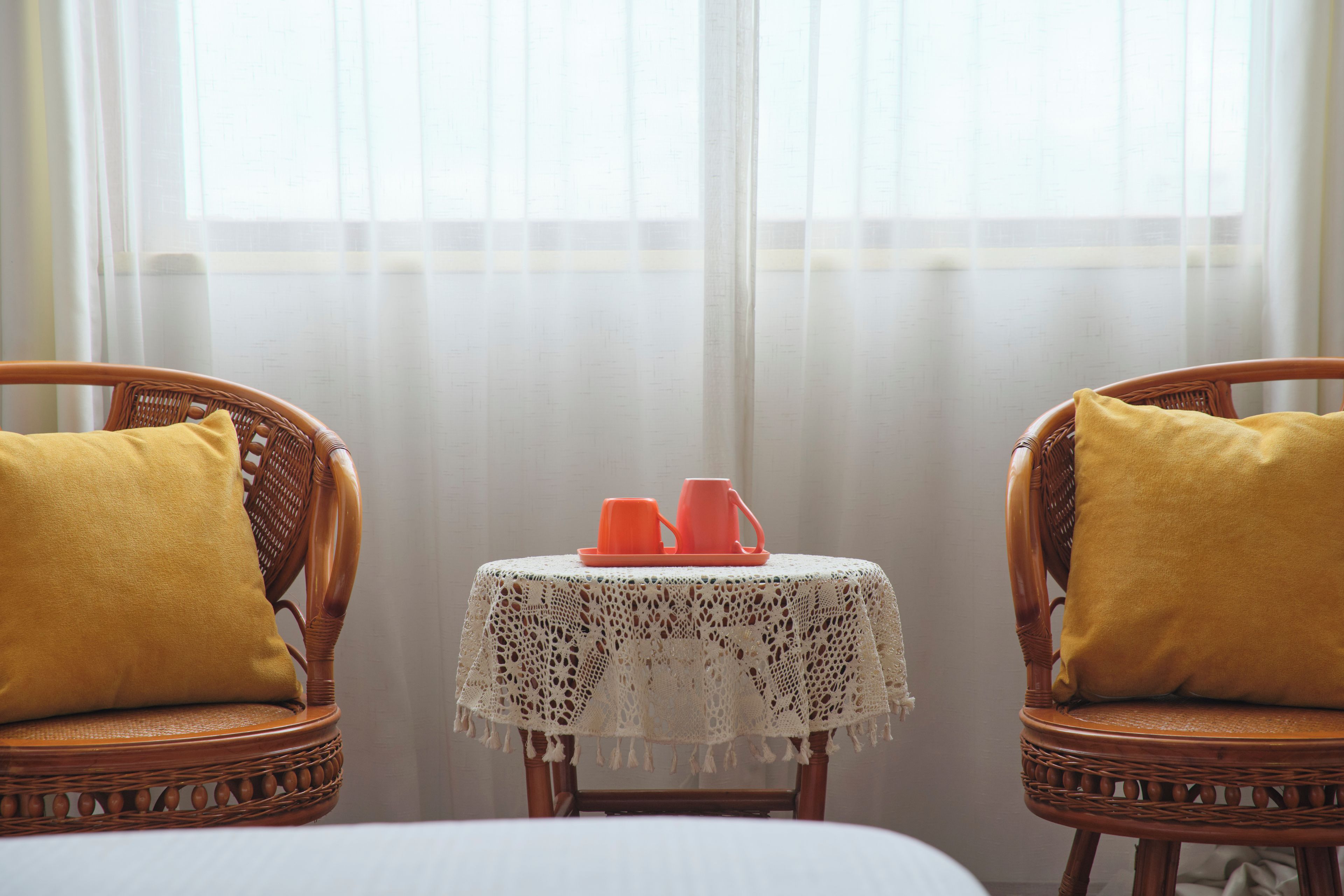 A room with two wicker chairs, a small table with a lace tablecloth, and a window with sheer curtains.