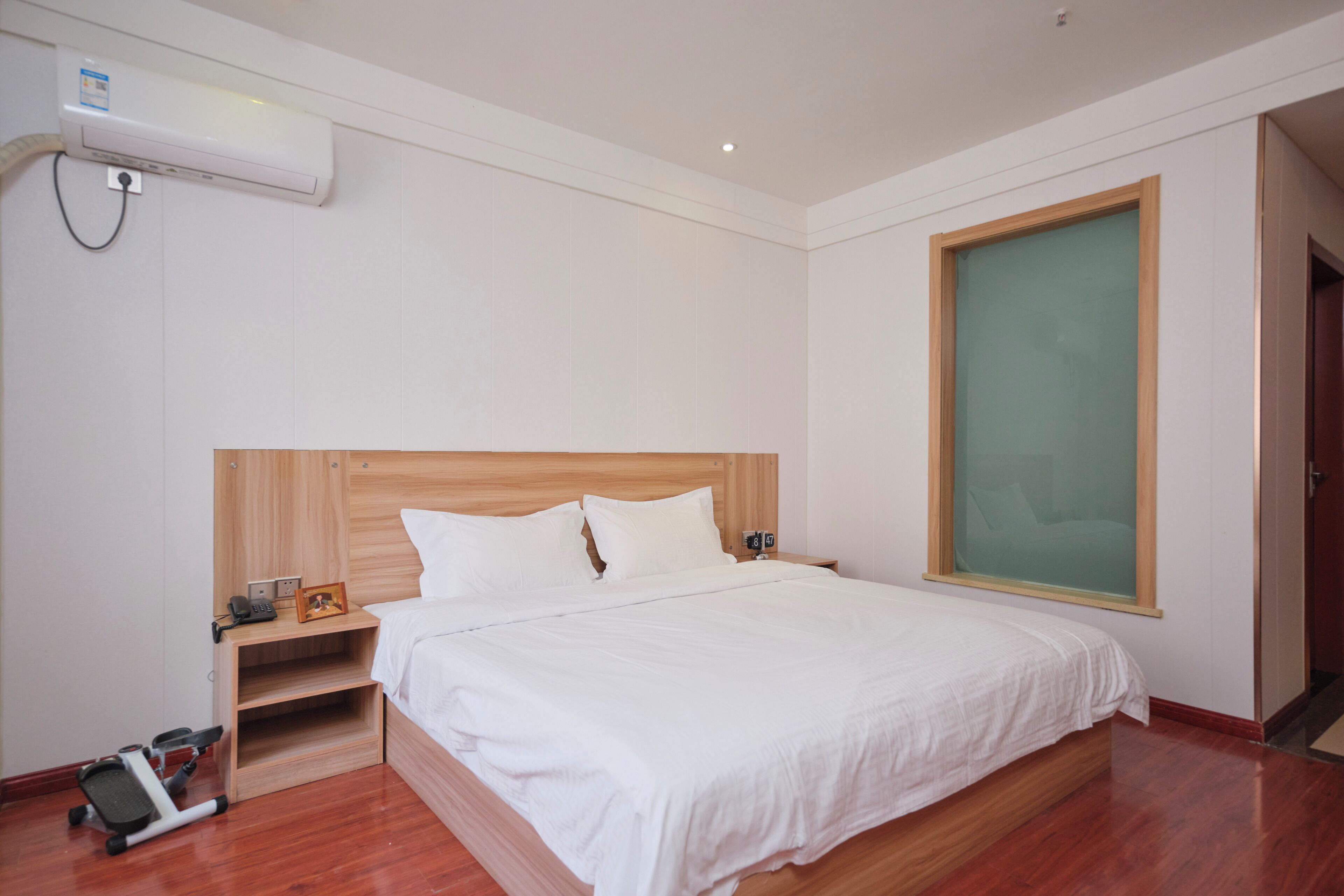A hotel room with a wooden bed, white bedding, a small bedside table with a lamp, a wall-mounted air conditioner, and a large window.