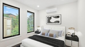 A bedroom with a large bed, two bedside tables, a wall-mounted air conditioner, and a window with a view of greenery. - French home walking to shops and shopping center (Maidstone)