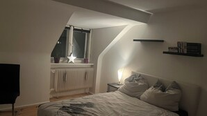 Ett sovrum med ett sluttande tak, ett element, ett nattduksbord med en lampa, en säng med rutigt sänglinne och ett fönster med växter.