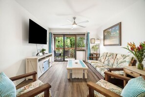 A living room with a TV, a ceiling fan, a sofa, a coffee table, and a view through a glass door. - Just Listed! Banyan Retreat at Kalapaki Bay Beach (Lihue)