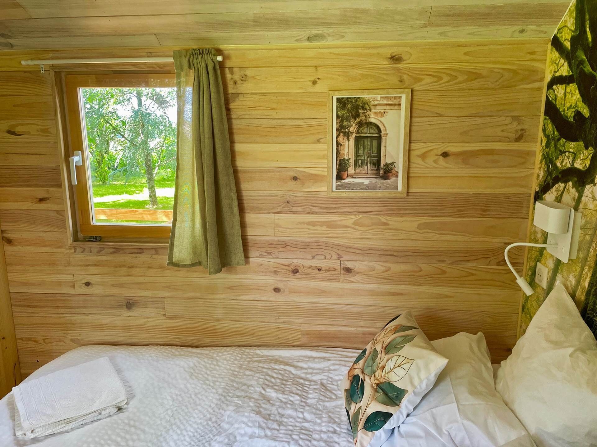 Une pièce de cabane en bois comprenant un lit, une fenêtre avec des rideaux, une lampe fixée au mur et un tableau encadré.