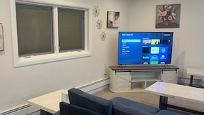 Uma sala de estar com uma grande televisão de tela plana, uma janela com persianas, um sofá e uma mesa pequena.