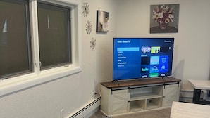 Uma sala de estar com televisão de tela plana, uma janela, uma mesinha e um sofá.