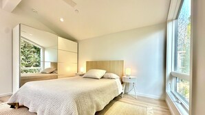 A modern bedroom with a large bed, a bedside table with a lamp, a large mirror, and a view of greenery outside. - Cozy 1-bedroom beach bungalow in beautiful Encinitas 1 block from surf with AC (Encinitas)