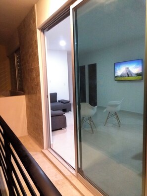 Ein modernes Interieur mit Glastür zu einem Wohnbereich, der eine Couch, Sessel und einen Fernseher zeigt, auf dem eine Landschaft zu sehen ist.
