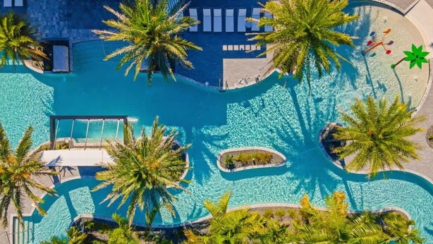 Luftaufnahme von Poolbereich eines Resorts mit Palmen, Liegestühlen und einem klaren, blauen Pool.