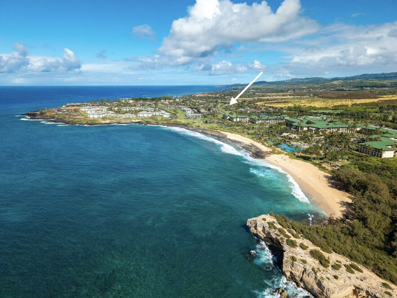 An aerial view of a coastal resort with a beachfront, clear blue waters, and a hilly landscape in the background.