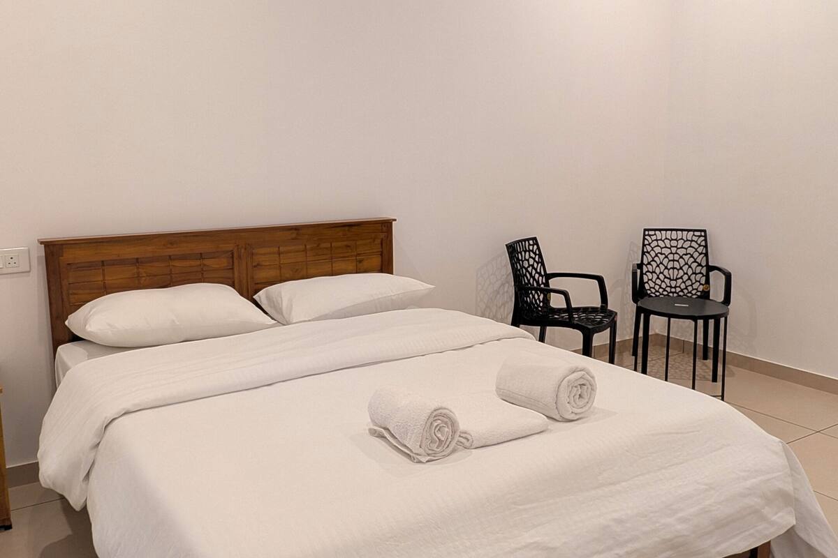 A neatly made bed with white linens and towels, a wooden headboard, and two black chairs with a geometric design.