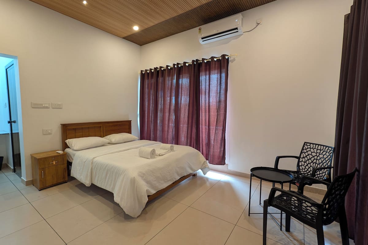A bedroom with a bed, a chair, a nightstand, an air conditioner, and a window with curtains.