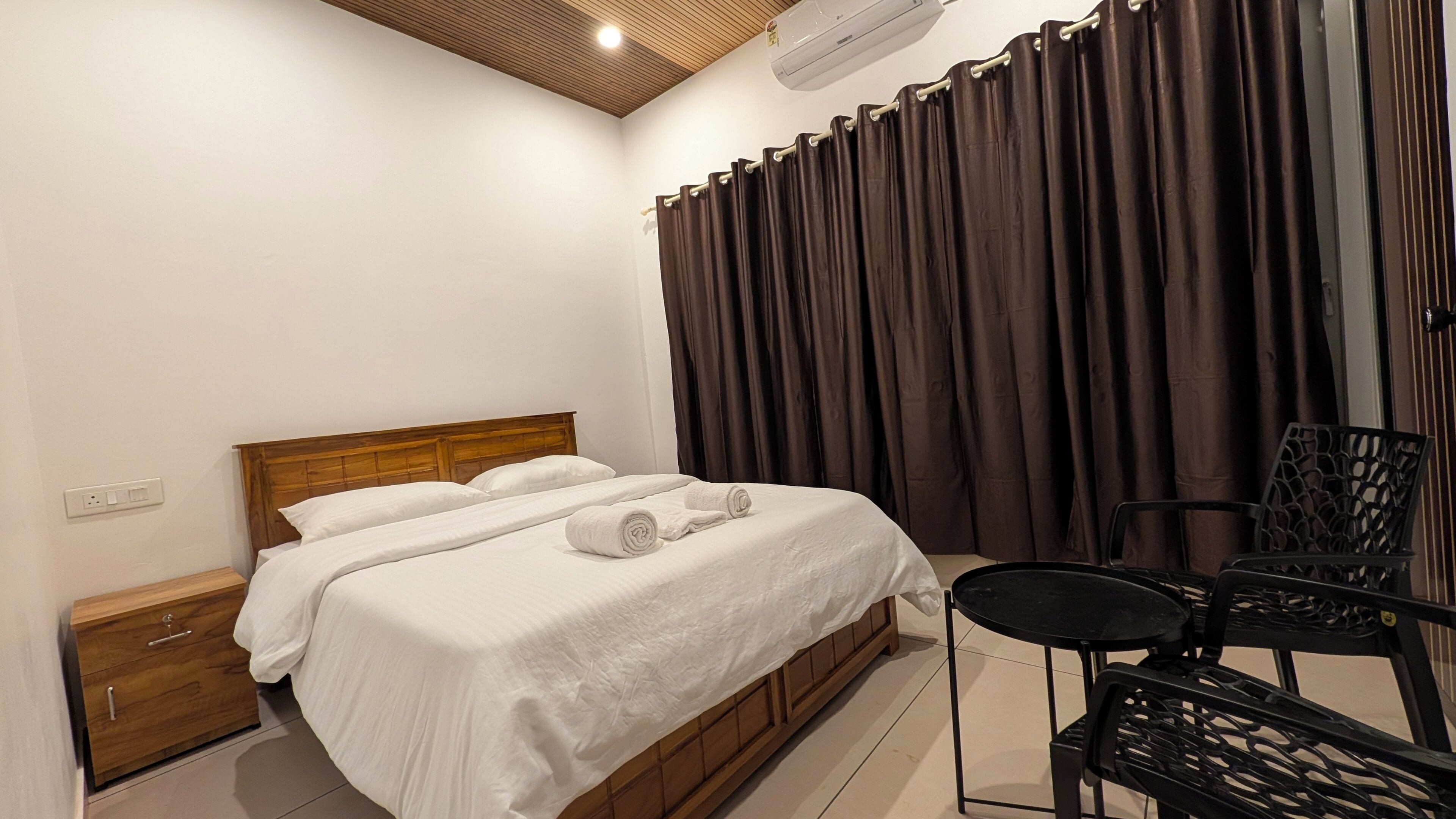 A bedroom with a wooden bed, a nightstand, a chair, and a large window with curtains.