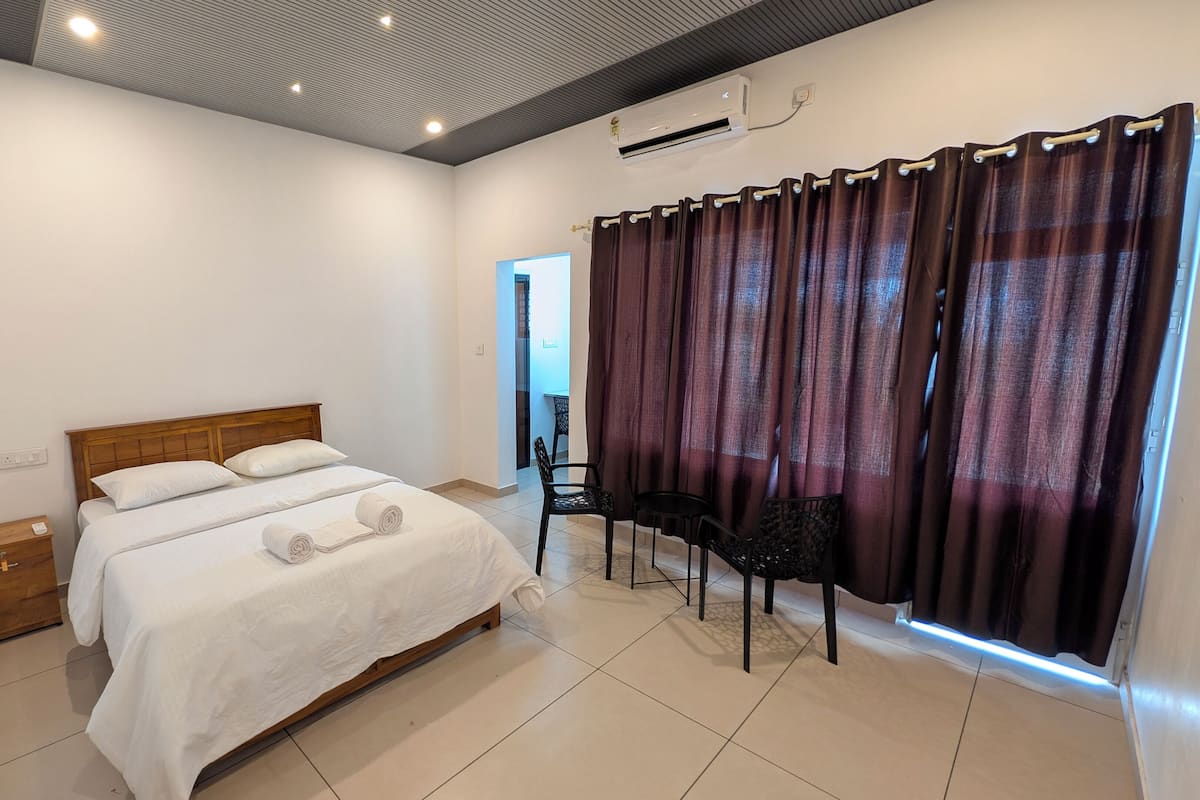 A hotel room with a wooden bed, a desk with two chairs, a window with curtains, and an air conditioning unit on the wall.