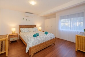 Quarto com cama de madeira, duas mesinhas de cabeceira, janela com cortinas leves e luminária no teto.