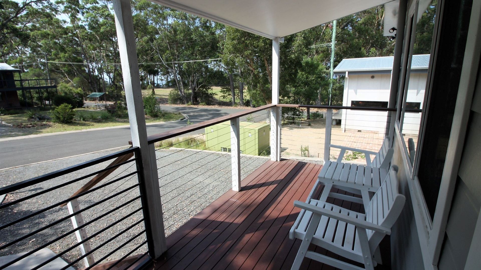 A covered porch with a wooden deck, white outdoor furniture, and a view of a road and greenery.