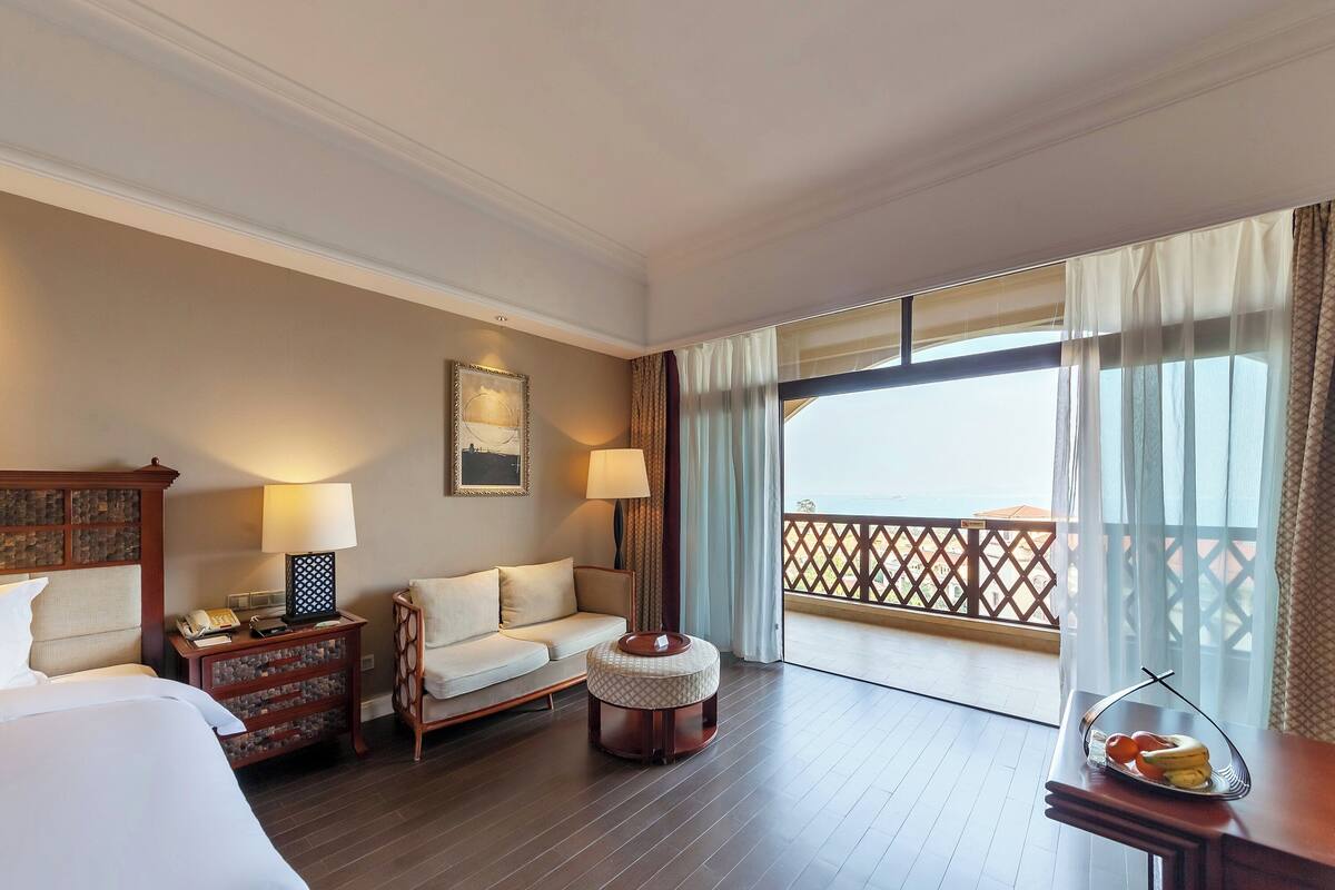 Habitación de hotel con cama, sofá, otomana redonda pequeña, mesita de noche con lámpara, un cuadro enmarcado y vista al balcón.