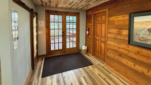 A hallway with wooden floors, a black doormat, a wooden door with glass panels, and a framed picture on the wall. - Rustline Haus - Cabin w/ Hot Tub, Walk to Village (Ruidoso)