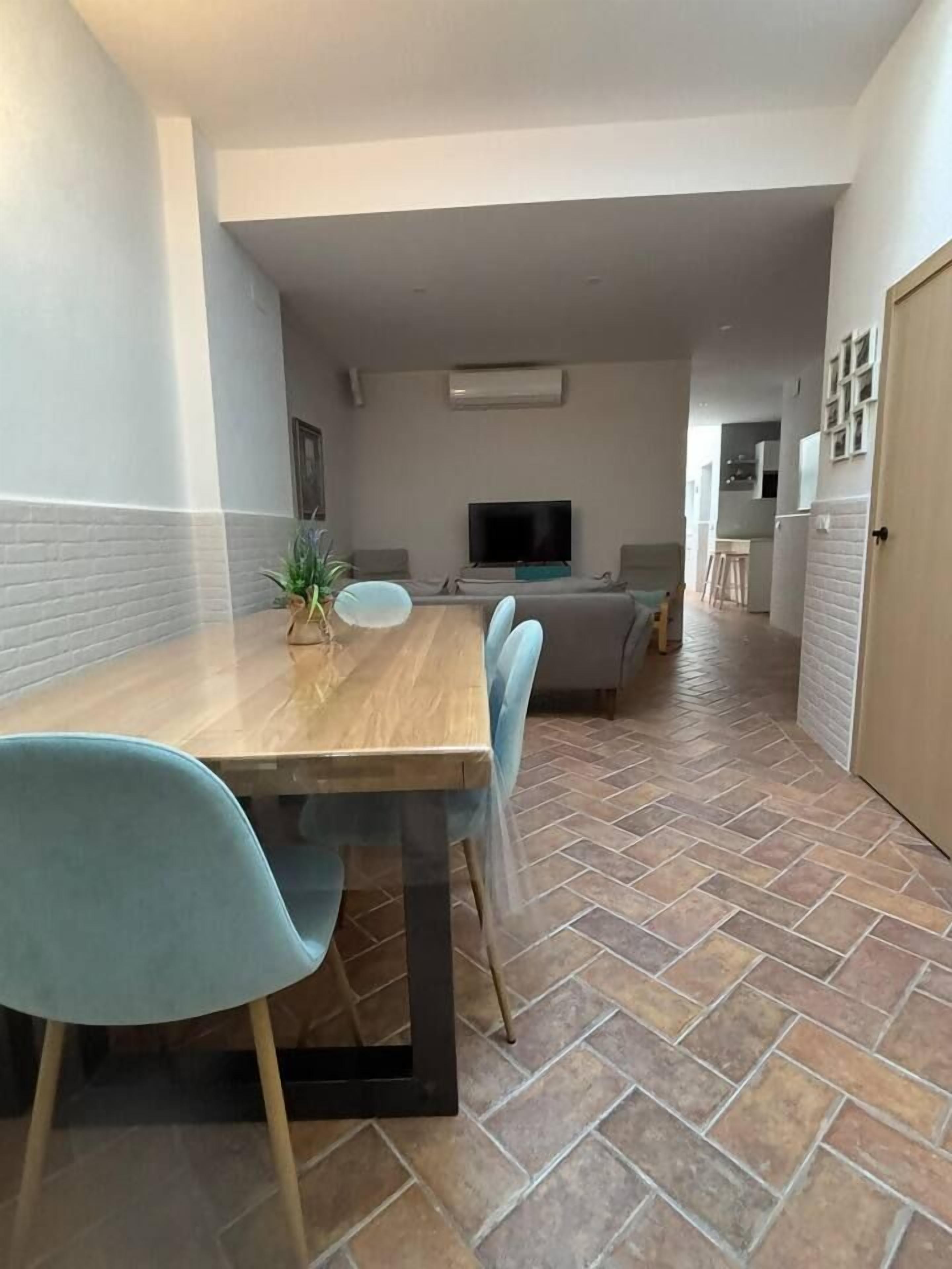 Un área de comedor con una mesa y sillas de madera, un televisor y una cocina al fondo.