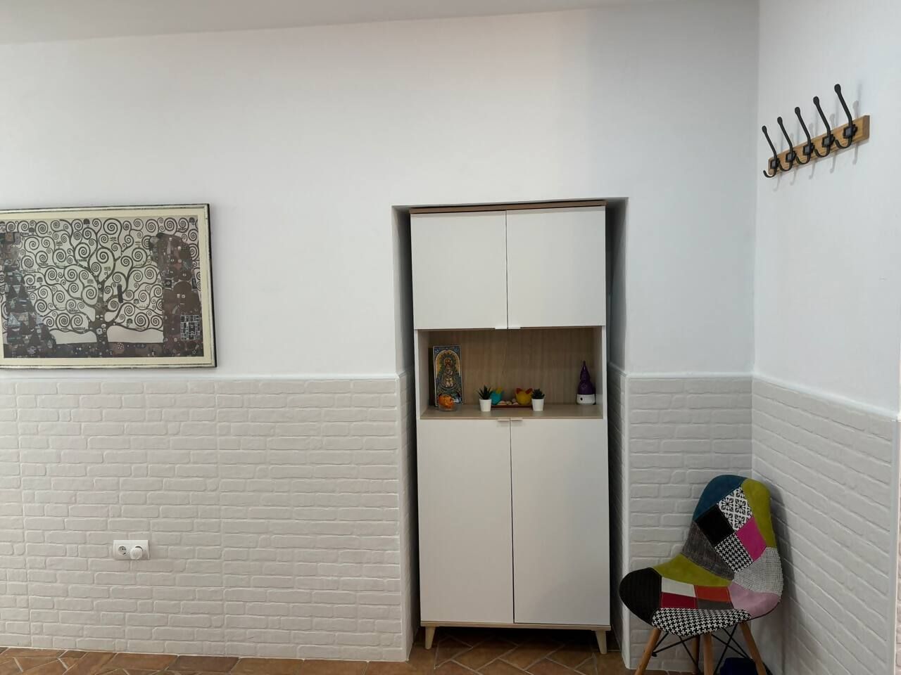 Una habitación con pared de ladrillo blanco, un armario blanco con espejo, una pequeña mesa con plantas y una silla colorida.
