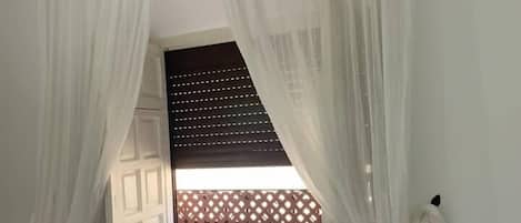 Ett rum med ett fönster som är täckt av tunna, vita gardiner, en trägallerstruktur som fungerar som avskärmning och en vit badrock som hänger på en krok.