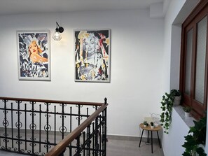 Un escalier orné d’une rampe en fer forgé, un mur avec deux affiches encadrées, et une petite table sur laquelle sont posés un panneau en forme de cœur et des plantes en pot.