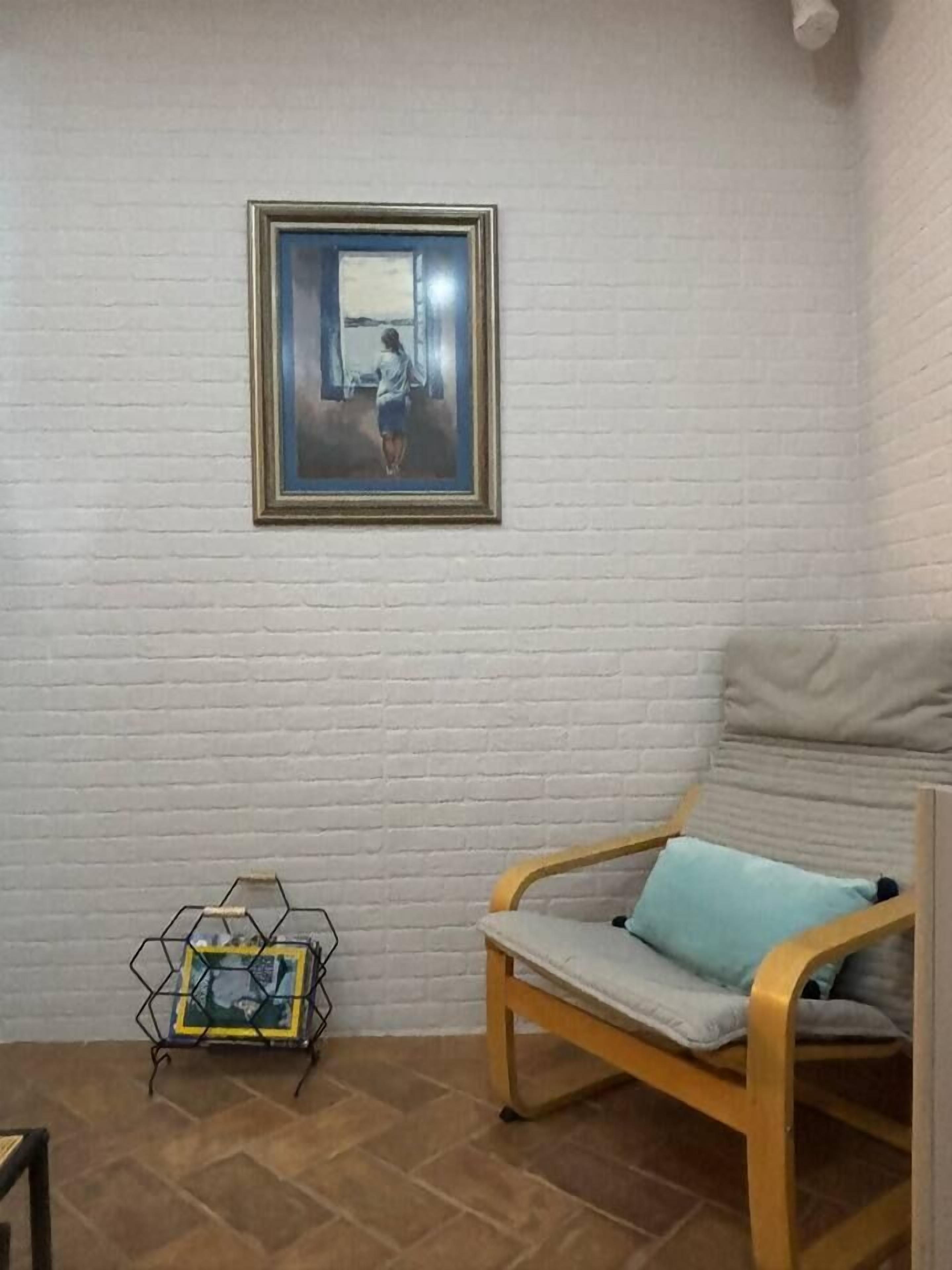 A room with a white brick wall, a framed painting, a wooden chair with a cushion, and a metal stand with a framed picture.