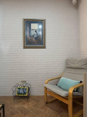 Une pièce avec un mur en briques blanches, un tableau encadré, une chaise en bois avec un coussin et un présentoir en métal avec une photographie encadrée.