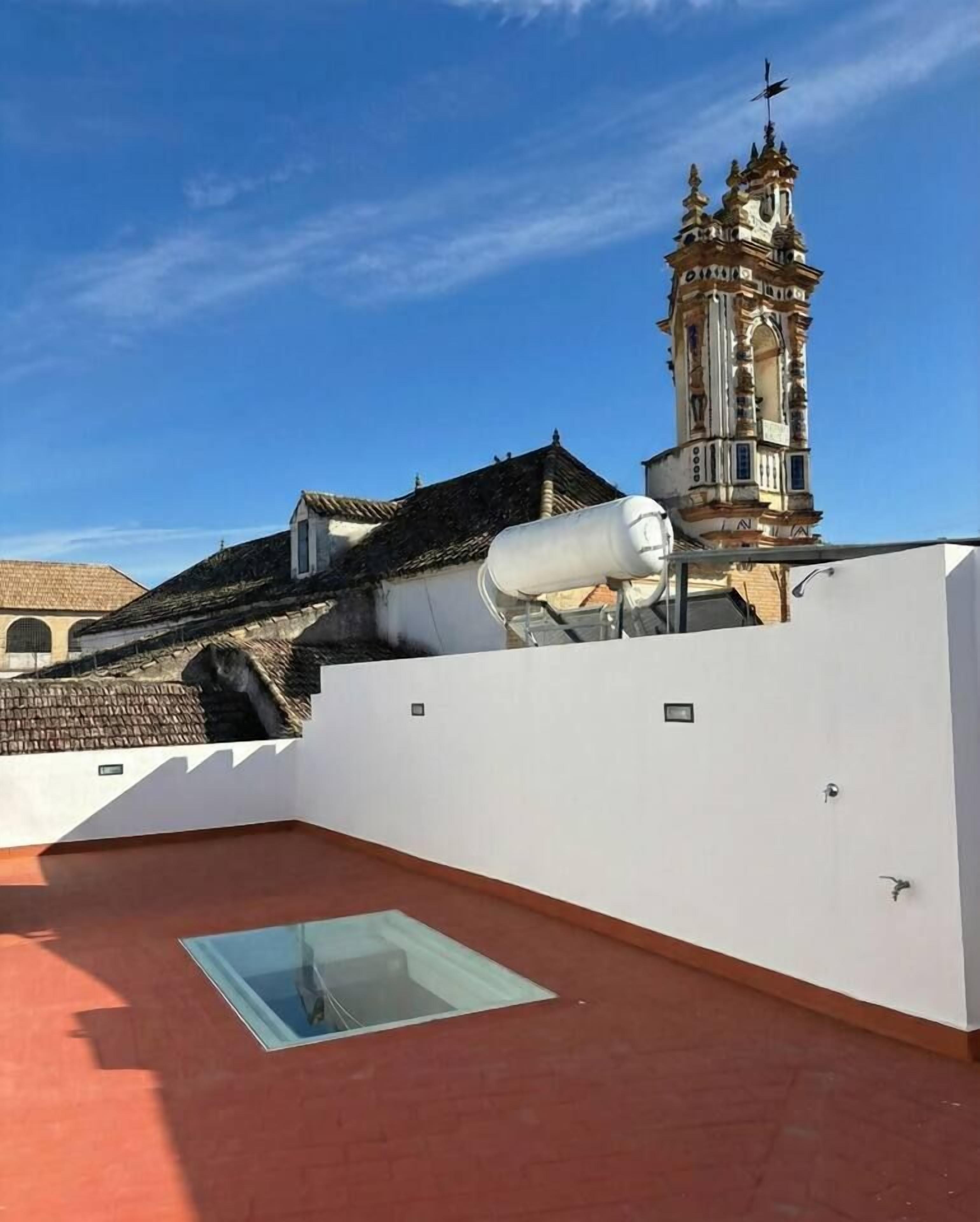 Une terrasse sur le toit, dotée d’une structure vitrée, d’un mur blanc et offrant une vue sur un bâtiment historique avec un clocher.