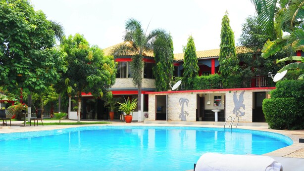 Ein Swimmingpool mit Liegestühlen, umgeben von tropischer Vegetation und einem Gebäude mit rotem Dach und Steinmauern.