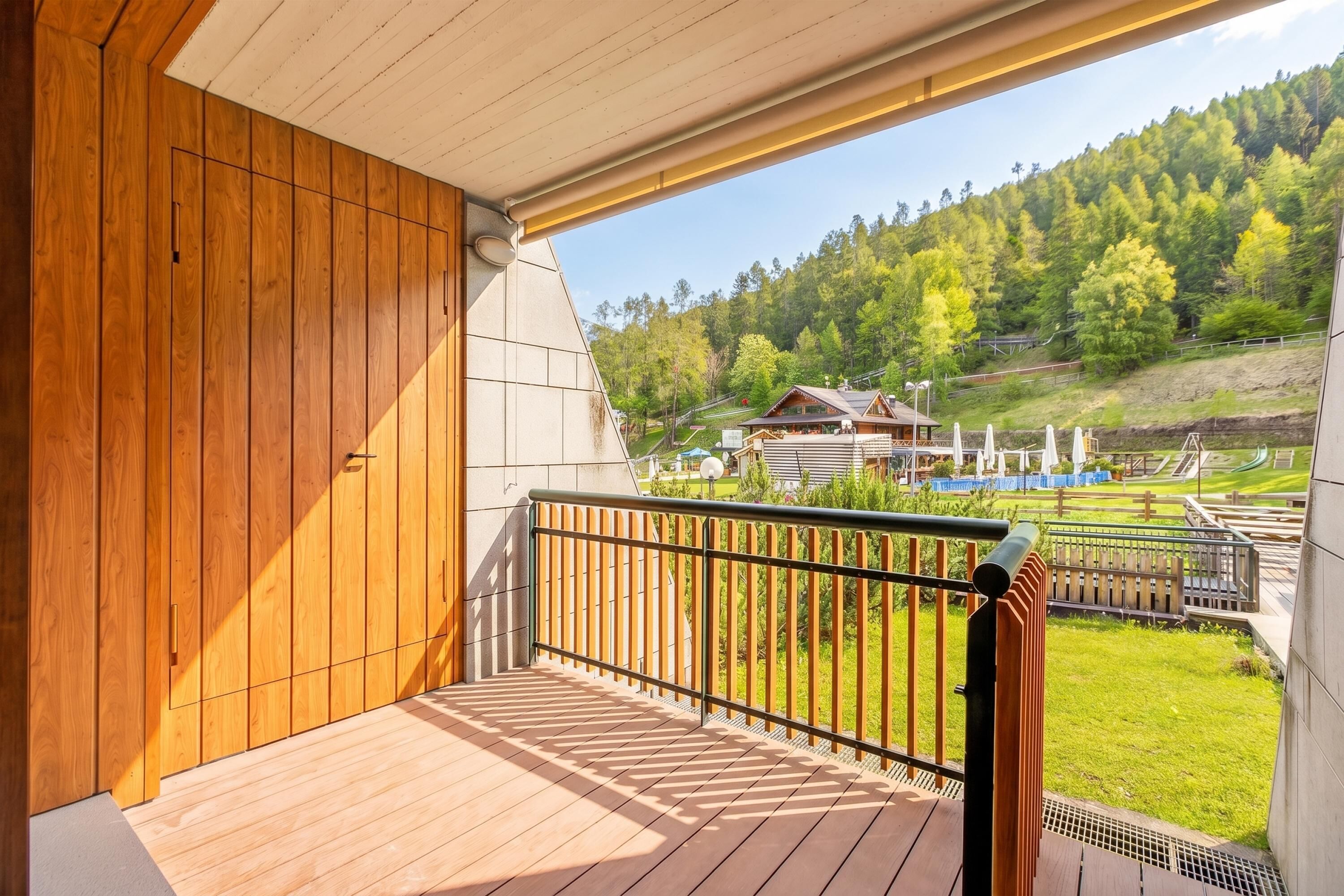 A wooden balcony with a metal railing, overlooking a green landscape and a building with a sloped roof.