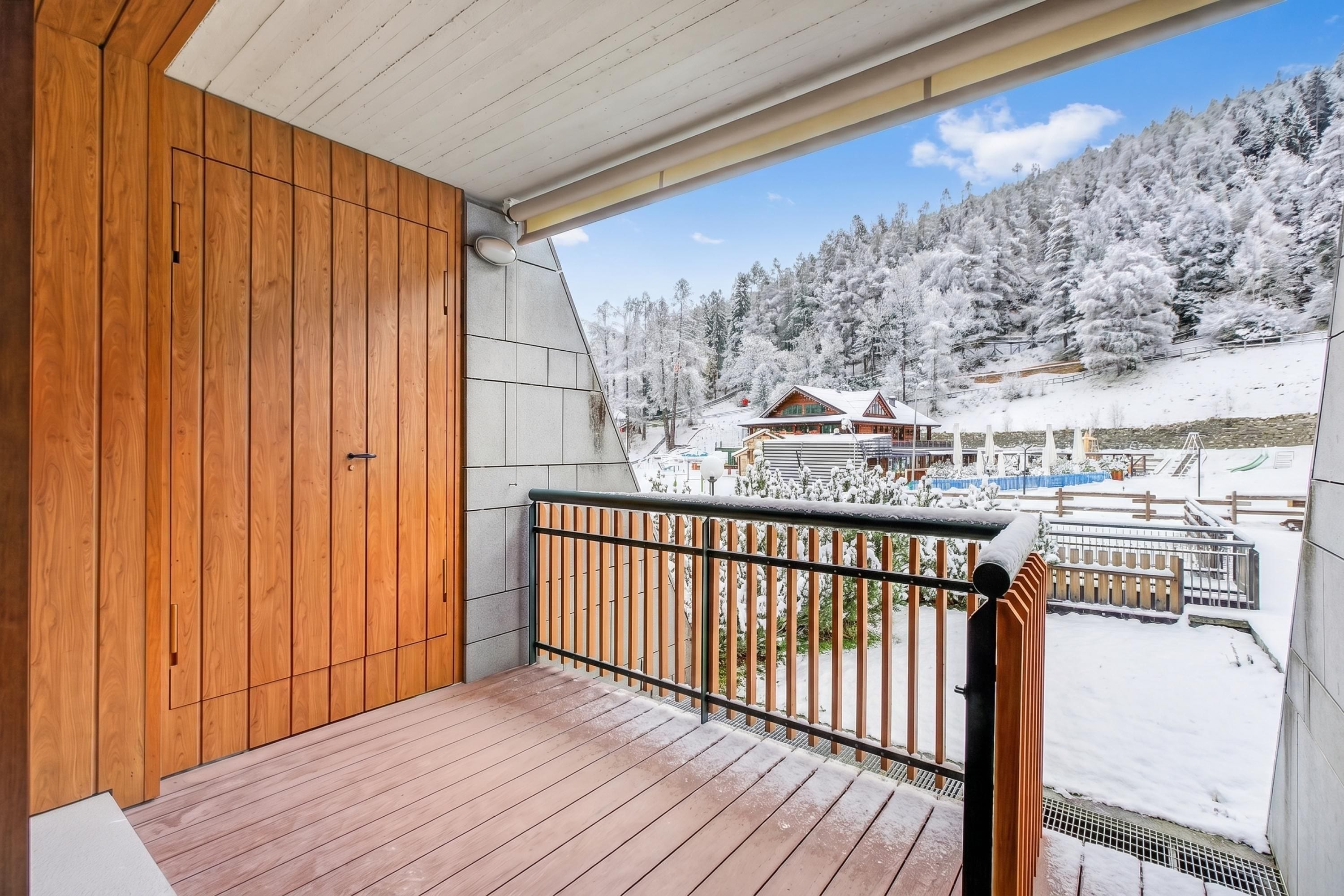 A wooden balcony with a metal railing overlooking a snowy landscape with buildings.