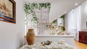 Ein Zimmer mit einem weißen Sofa, einem Holztischchen mit Vase und Kerzen, einer dekorativen weißen Trennwand mit Grünpflanzen und einem Fenster mit Vorhängen.
