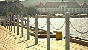 Um deck de madeira com mourões brancos e uma barreira de corrente, com vista para um corpo de água e edifícios ao fundo.