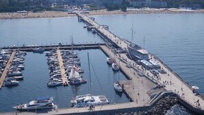 Uma marina com vários barcos atracados, um cais com pessoas a passear e uma praia ao fundo.