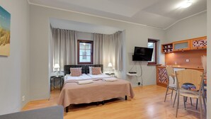 Quarto de hotel com cama, mesas de cabeceira, televisão, escrivaninha com cadeiras e cozinha.